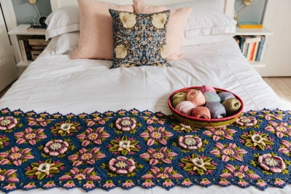 785891265 Cozy bedroom scene with a white bed, pink and patterned throw pillows, and a navy crochet floral runner along the foot of the bed, plus a bowl of colorful yarns on top edge of the runner.