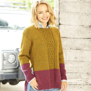 Smiling woman wearing a mustard-yellow and burgundy color-block knit sweater and jeans, standing beside a wooden wall outdoors.