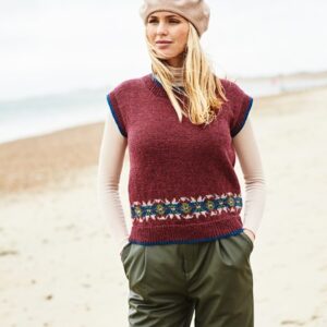 Woman on a sandy beach wearing a beige beret, burgundy knit vest with a patterned band, and olive pants with hands in pockets.