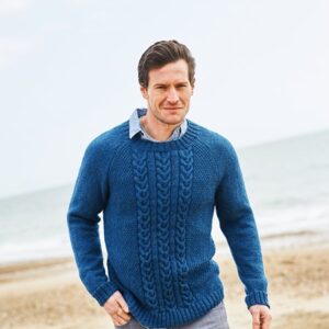 Man walking on a sandy beach in a blue cable-knit sweater over a light collared shirt.