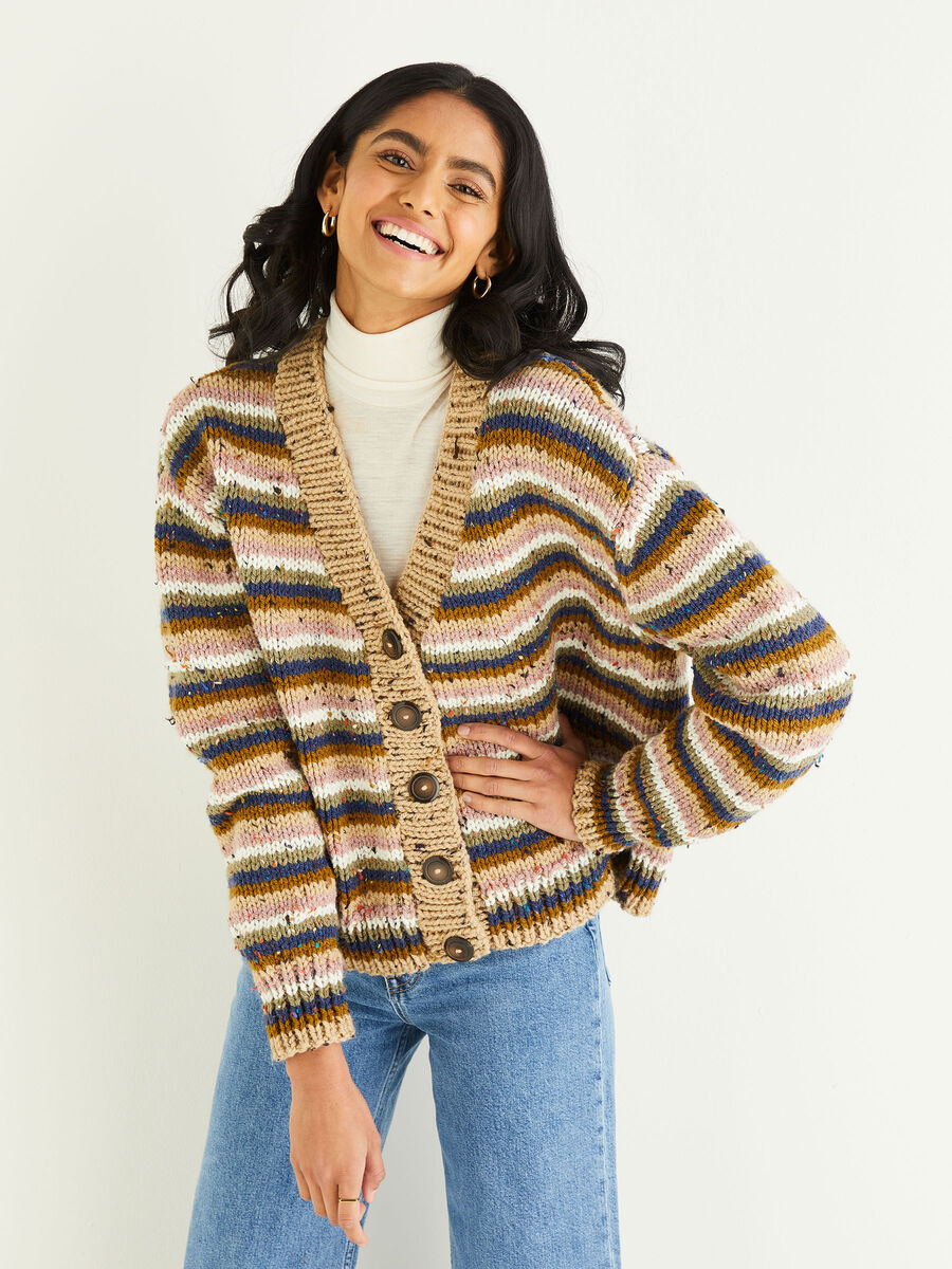 Woman smiling in a chunky multicolored striped cardigan over a cream turtleneck and blue jeans.
