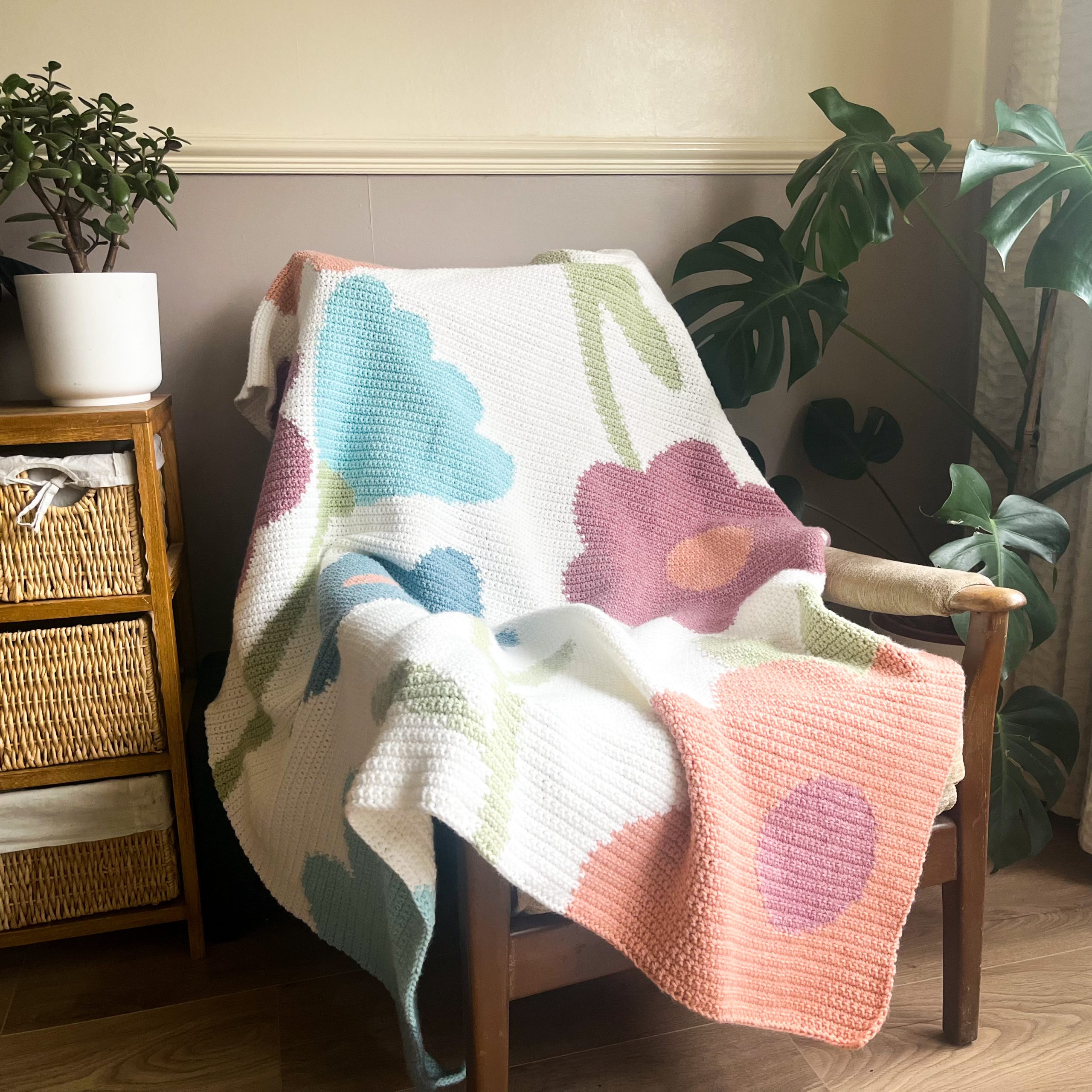 IMG_8792_jpg-scaled Wooden chair with a pastel knitted blanket draped over it, a white pot with a small plant on a side table, and large leafy houseplants nearby in a cozy indoor scene
