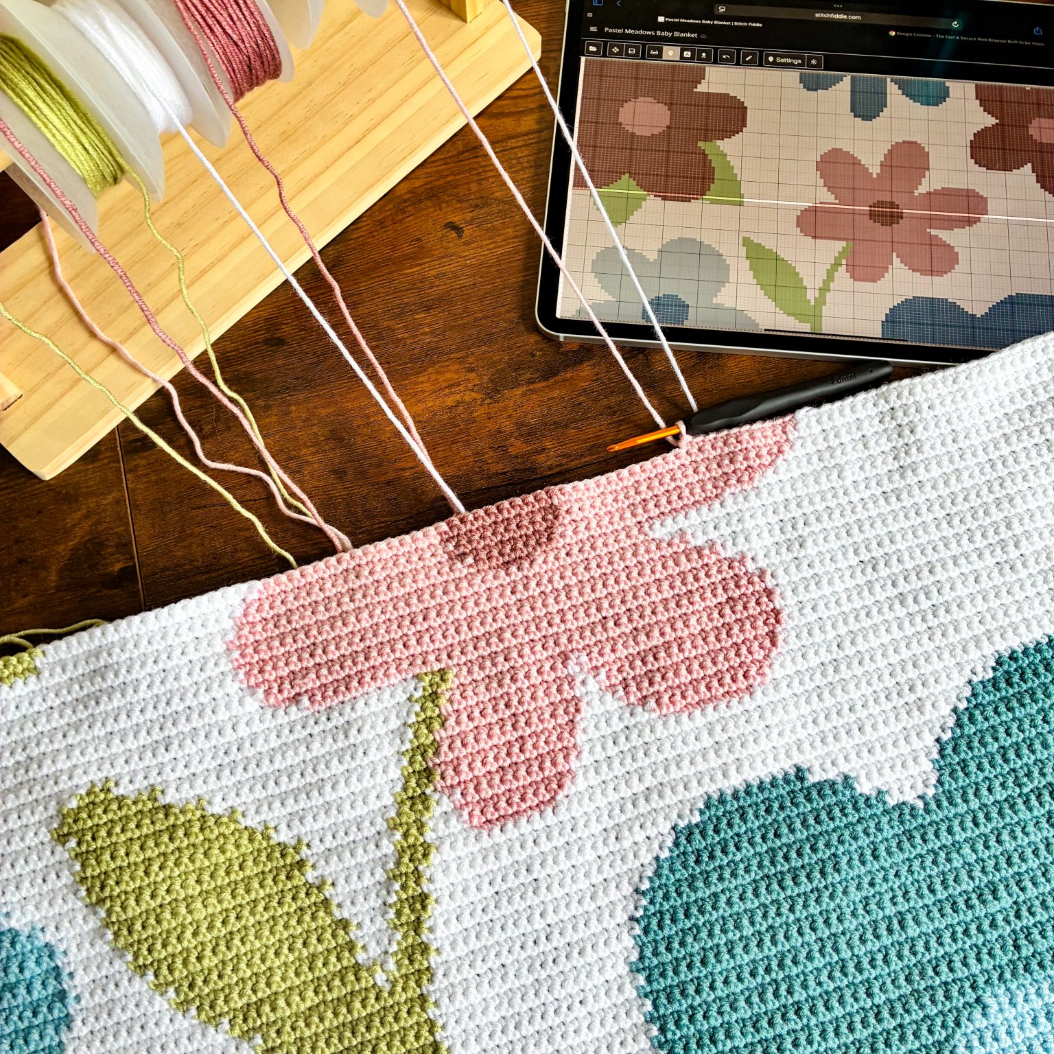 Pastel-Meadows-Baby-Blanket-WIP-1-1536x1536 (1) Top-down view of a crochet workspace: pastel yarn skeins on a rack, a pink-and-white crocheted blanket in progress, a crochet hook resting on the fabric, and a tablet displaying a floral grid pattern.