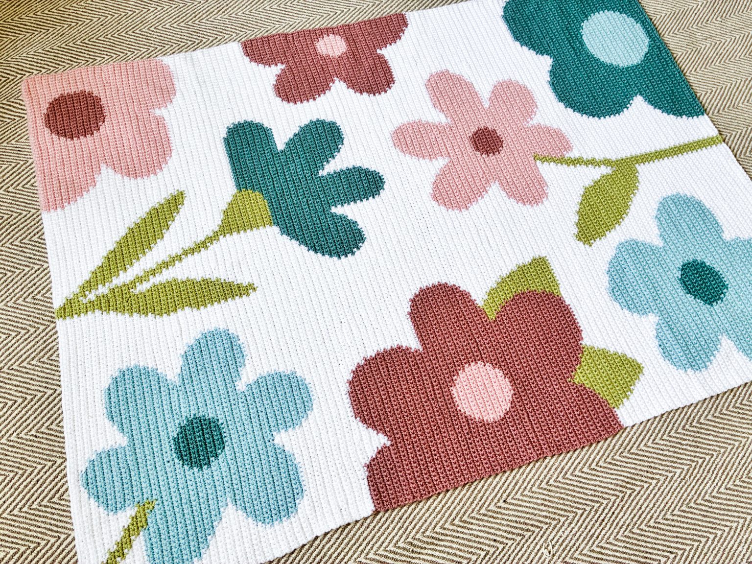 pastel-meadows-floral-crochet-blanket-1536x1152 Colorful floral rug with large pink, red, teal, blue, and green flowers on a white background, laid on a herringbone floor.