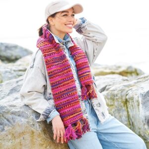 Smiling woman in a denim jacket and jeans leaning on rocks at the coast, wearing a pink striped scarf and beige cap.