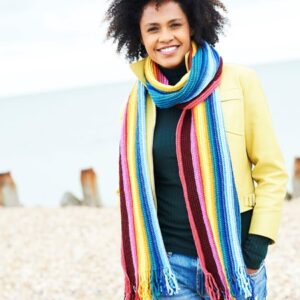 Smiling woman in a yellow coat and colorful striped scarf standing on a pebble beach near the sea.