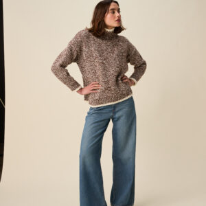 10948 Mingle High Neck Sweater img1Person standing with hands on hips, wearing a chunky brown-and-cream knit sweater, light-wash jeans, and red sneakers against a beige studio backdrop.