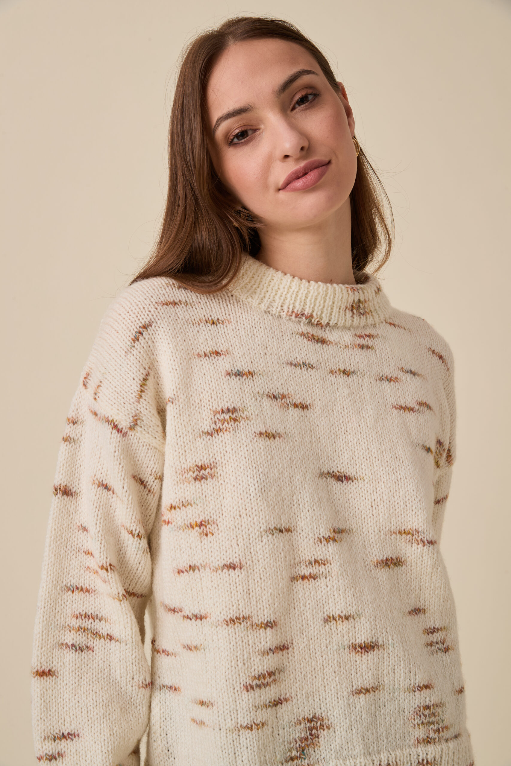 10958 Bright Sparks Sweater img2 Close-up portrait of a young woman with light makeup wearing a cream knitted sweater against a beige background.