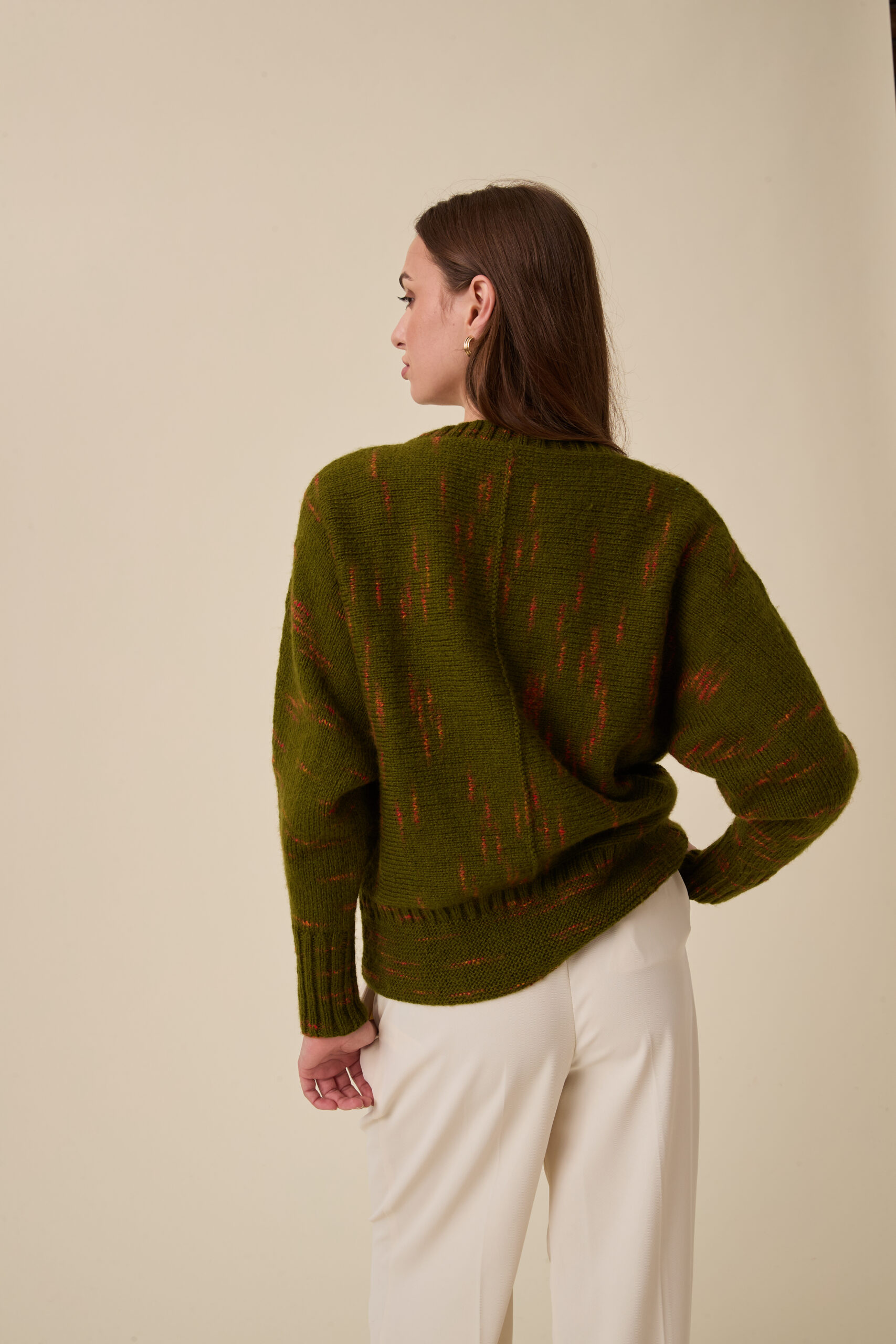 10963 Glimmer Sideways Sweater img3 Woman seen from the back with her head in profile, wearing an olive-green knitted sweater with orange flecks and white pants against a beige backdrop