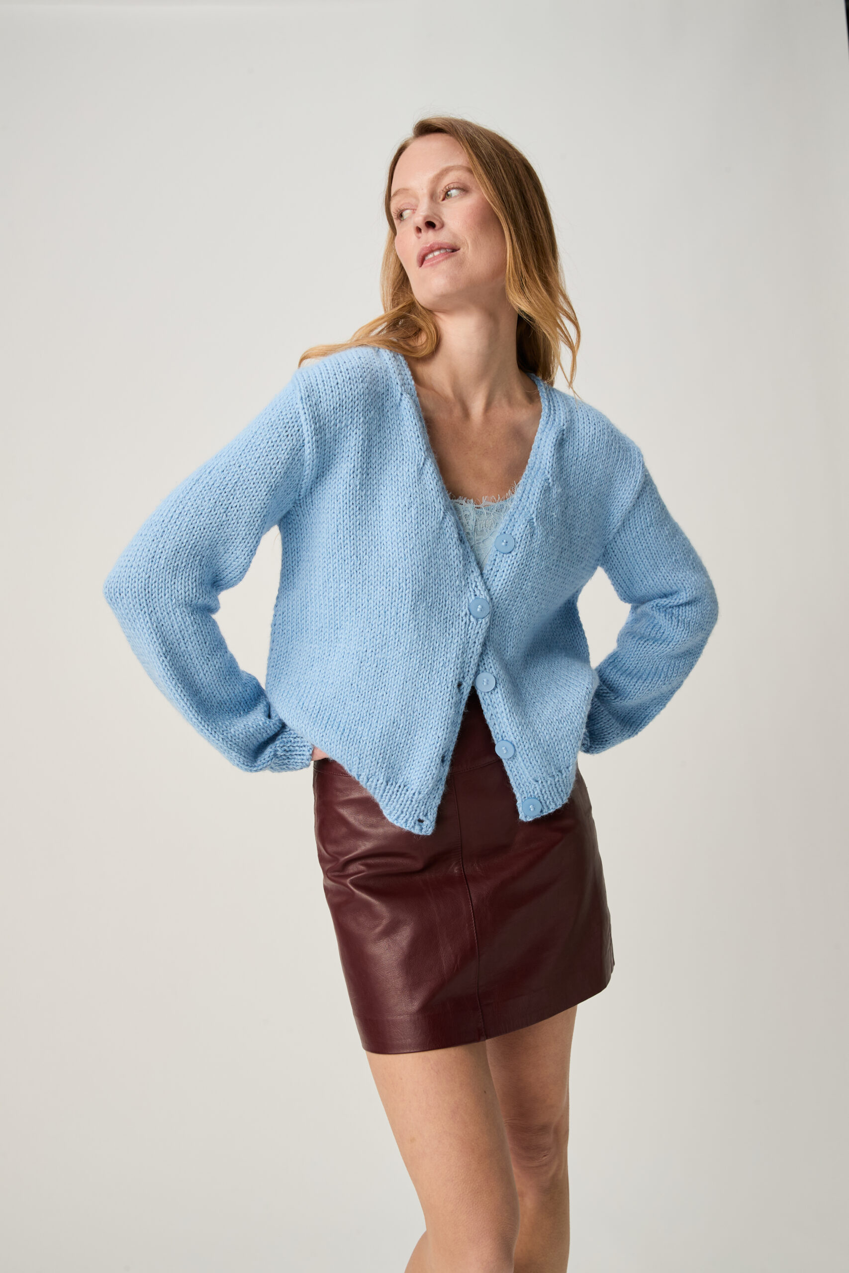 Woman posing in a light blue knitted cardigan over a pale top, burgundy leather skirt, looking to her left with hands on hips. 10967 Stem V Neck Cardigan img1