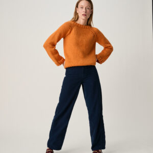 10972 Propagate Raglan Sweater img1Full-body shot of a woman standing with hands on hips, wearing an orange knit sweater, dark blue pants, and burgundy shoes in a studio.
