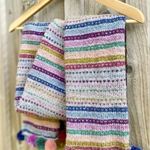 Colorful striped knit scarf with multicolor pom-poms hanging on a wooden hanger against a fence.