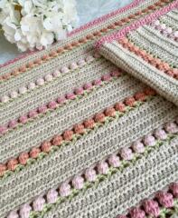 Crocheted striped blanket in pastel beige, pink, and green laid out with white flowers in the corner.