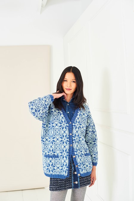 Young woman wearing a blue geometric-patterned cardigan over a denim shirt in a bright white room.